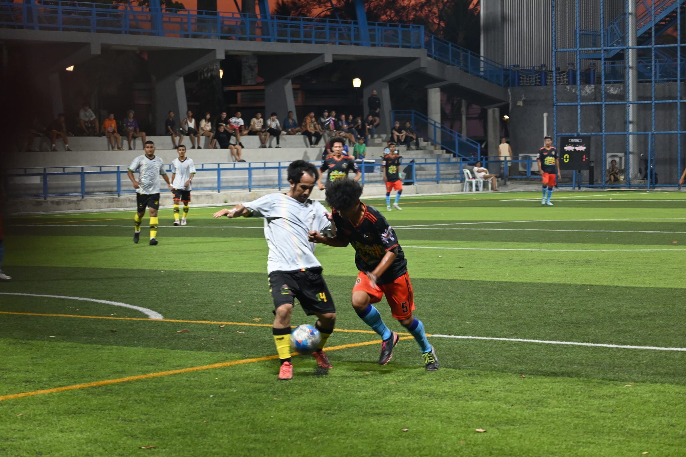 Evening match under floodlights: a player in white controls the ball as an opponent in navy approaches.